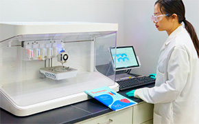 A laboratory benchtop bioprinting system is shown inside a clean research environment. The enclosed instrument features multiple printheads positioned above a printing stage within a transparent chamber. A researcher in a laboratory coat is seated beside the instrument, operating the system through a connected computer workstation.