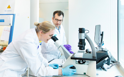 HUB Organoids scientists at the microscope A smiling female scientist wearing gloves looks into a microscope while a male wearing glasses and purple gloves assists at the computer. HUB Organoids logo partially appears on a lab coat.