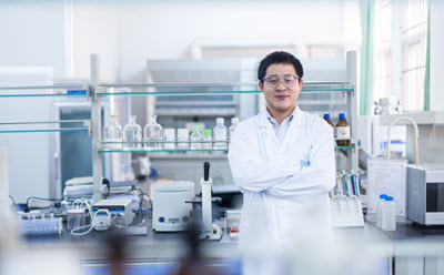 Pharma & Biopharma Manufacturing Scientist in pharma lab in a white lab coat stands with arms crossed in a laboratory setting, surrounded by various scientific equipment and glassware.