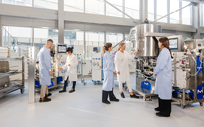 Researchers working around stainless steel lab equipment Researchers in white lab coats and hairnets work around stainless-steel processing equipment in a bright laboratory