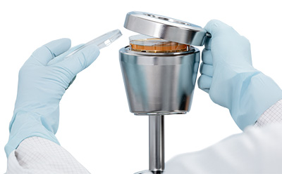 A close-up of a magnetic stainless steel lid with a handle being removed from a microbial air sampler, showing a 90 mm settle plate inside.