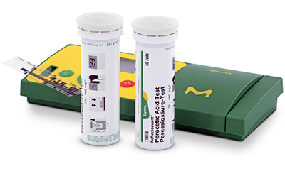 Two cylindrical containers of diagnostic test strips positioned next to a green box labeled with a yellow and green design and medical symbols. The containers have labels providing information about the test strips inside, and one container is open with a strip partially extended, suggesting readiness for use in a clinical or laboratory setting.