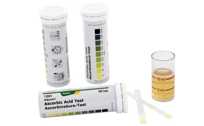 Three containers of test strips, each labeled for different chemical tests including an ascorbic acid test. Two of the containers are closed with labels showing color scales and instructions, and one is open with several test strips extended. Additionally, there is a small glass beaker with a yellowish solution, suggesting a setup for conducting these tests.