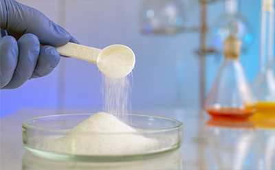 Carbohydrate powder Gloved hand adding white powder to a glass beaker in a laboratory, with colorful flasks and scientific equipment in the background.