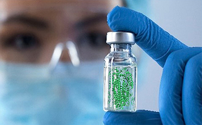 Green glycoconjugate beads Gloved scientist holds a vial containing green particles, with a masked laboratory worker blurred in the background.