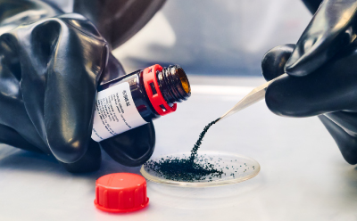The image shows a close-up of a laboratory setting where an individual, wearing black gloves, is transferring a fine black powder from a bottle into a petri dish using a spatula.