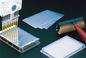 将液体注入孔板的多通道移液器，以及旁边的 AlumaSeal 96 ® 箔、一叠滤纸和一个滚瓶，都放在黑色背景的暗面上。