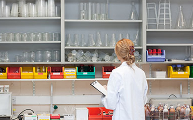 Lab worker looking at inventory on shelves 一个身穿白大褂的人站在实验室里,面对镜头,手里拿着一个写字板。实验室的架子上摆放着各种实验设备,如烧杯、烧瓶、试管和装有不同颜色化学试剂的瓶子。人物前面的长凳上还摆放着试管架和其他科学仪器。这样的布置让人联想到一个专业的研究环境,突出了科学实验中使用的精心组织和各种工具。