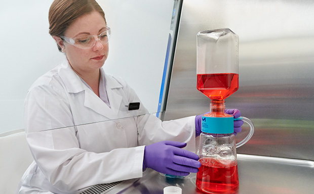 Sterile Filtration Devices 一位身着白大褂的女士正在操作装有红色液体的实验室烧瓶,烧瓶与另一个底部装有蓝色液体的设备相连。