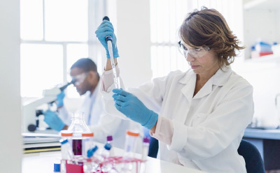 Scientist performing liquid handling with a pipette in a laboratory, with another scientist working at a microscope in the background. Scientist pipetting a sample at a lab bench while another scientist uses a microscope in the background.