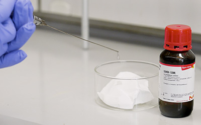 Handling demonstration of non pyrophoric organolithium solution during dispensing A gloved hand holds a syringe releasing a small droplet above a clear glass dish, with a labelled amber bottle of organolithium solution placed nearby on the laboratory benchtop.