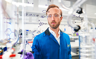 Chemist documenting reaction schemes on a transparent board within a modern laboratory environment. A laboratory scientist in a blue lab coat writes chemical structures on a transparent glass surface, surrounded by lab equipment and instrumentation in a bright, well organized research space.