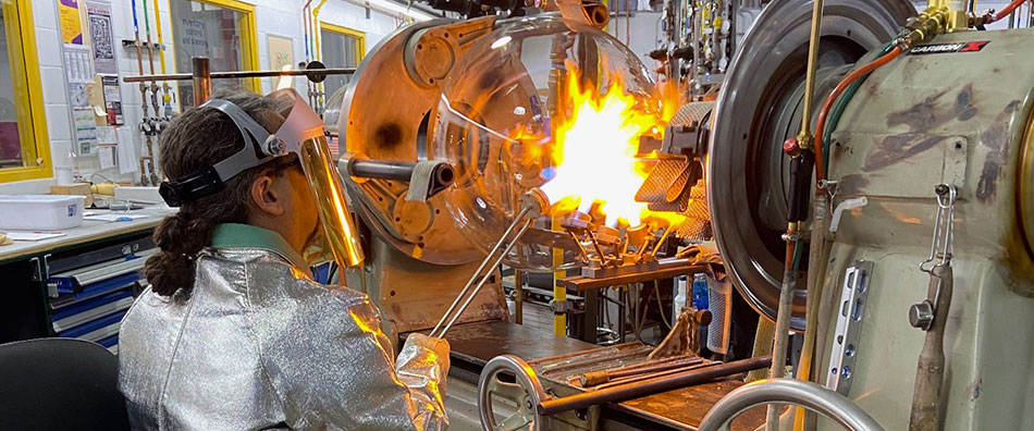 Glassblower works on flask 圆底烧瓶位于车床中,玻璃技师将火焰引至烧瓶颈部。玻璃技师身着防护服,包括专用外套、手套和面罩。