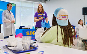 A child wearing a "Thinking Cap" hat is seated at a table, facing a presentation about water filtration, with two adults standing in front of a large screen displaying relevant information. The adults are engaged in explaining the topic, while various educational materials, including cups and worksheets, are visible on the table, creating an interactive learning environment.