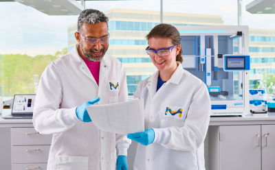 Scientists with Paper In Front of AAW™ Workstation 2 scientists standing in front of AAW™ automated assay workstation holding paper and chatting to each other.