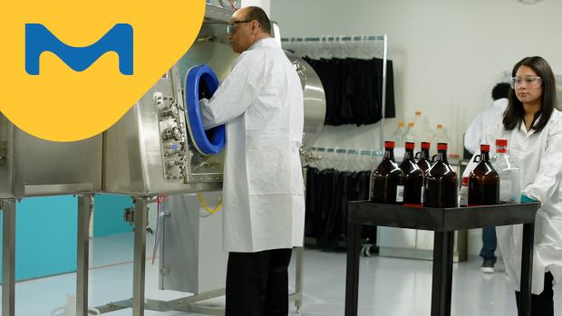 <p>HPAPI生产成功的秘诀</p> A male scientist in a lab coat is using an isolator, while a colleague female scientist in a lab coat is transferring chemicals into the training facility behind him.
