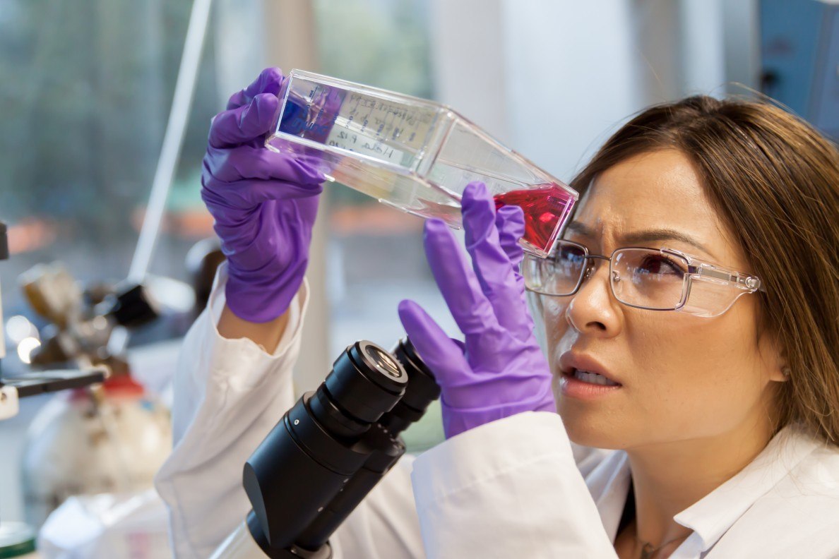 A scientist in a lab coat and purple gloves holding and examining a clear vial containing a red liquid. Laboratory equipment, including part of a microscope, is visible, indicating a scientific laboratory setting.