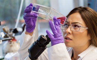 scientist in a lab coat and purple gloves holding and examining a clear vial containing a red liquid. Laboratory equipment, including part of a microscope, is visible, indicating a scientific laboratory setting