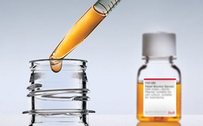 Serum bottle in the background halfway filled with golden liquid. In the foreground, a pipette partially filled with gold liquid is pulling out of a bottle top and dripping.