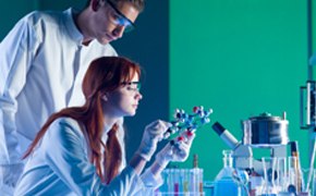 Two scientists are working in a laboratory with a green background. One scientist is observing a molecular model held by the other, who is focused on the model while wearing gloves. Various laboratory equipment, including flasks and a microscope, is visible on the table in front of them.