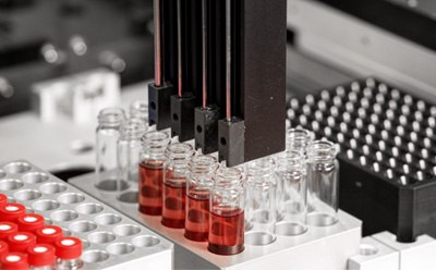 A close-up view of a laboratory automation system featuring multiple robotic arms positioned above several glass vials containing red liquid. The vials are arranged on a tray with circular openings, and the background includes various laboratory components and equipment.
