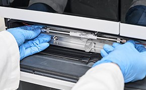 A scientist in a laboratory, wearing blue gloves, is inserting an HPLC column into a chromatography machine for analytical chromatography.