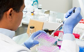 A scientist in a laboratory, wearing a white lab coat, glasses, and blue gloves, is working with biochemicals. He is using a pipette to transfer a pink solution into a multi-well plate. The lab bench is filled with various lab equipment and containers, indicative of an active biochemical research environment.