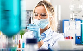 A scientist in a laboratory environment, wearing a white lab coat, safety glasses, a face mask, and blue gloves, is working with crosslinkers. She is using a pipette to handle a liquid sample, with various lab equipment and containers visible in the background. The scene depicts a focused and sterile research setting.