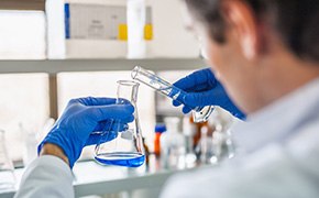 A scientist in a laboratory setting, wearing a white lab coat and blue gloves, is working with peptide synthesis reagents. He is carefully pouring a clear liquid from a test tube into an Erlenmeyer flask containing a blue solution. The background features shelves with various lab supplies and equipment, indicating an active research environment focused on peptide synthesis.