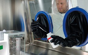 A scientist wearing a protective suit and working inside a fume hood, also known as a chemistry hood, which is a ventilated enclosure with a transparent front and built-in gloves, designed to safely contain and exhaust hazardous fumes, vapors, and dust during chemical experiments. The scientist is manipulating a container inside this controlled environment, ensuring safety and precision.