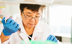 A scientist in a laboratory environment, wearing a white lab coat, glasses, and blue gloves, is using proximity labeling kits for micromapping. She is carefully handling a pipette and working with lab equipment, focusing intently on her task.