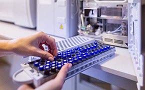 A tray of blue-capped vials are placed into a laboratory instrument, likely a chromatograph, for chemical analysis. The machine is partially open, revealing its internal components, indicating preparation for testing or calibration in a chemistry lab setting.