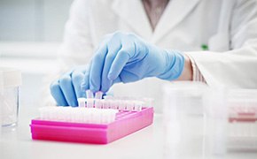 A scientist wearing a white lab coat and blue gloves is handling small test tubes arranged in a pink rack. The test tubes are part of Aldrich Market Select's small molecule libraries, used for the rapid screening of compounds. The background shows a bright, clean laboratory environment with additional laboratory supplies and containers visible, emphasizing the organized and methodical nature of scientific research and compound screening.