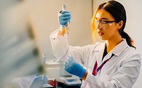 A scientist in a laboratory setting, wearing a white lab coat, safety goggles, and blue gloves, is working with PROTAC® protein degrader building blocks. She is handling a pipette and carefully transferring a liquid into a vial, demonstrating precision and focus in her research work.