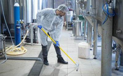 A worker in protective clothing is using a long-handled brush to clean the floor in a food processing facility. The background shows various equipment and tanks, emphasizing the importance of hygiene monitoring in food testing A worker in protective clothing is using a long-handled brush to clean the floor in a food processing facility. The background shows various equipment and tanks, emphasizing the importance of hygiene monitoring in food testing