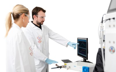 Two scientists in lab coats and gloves working with laboratory equipment. One is pointing at a computer screen displaying graphs, while the other looks on attentively. The scene takes place in a modern laboratory environment.