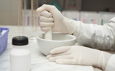 A close-up view of a gloved hand holding a pestle, grinding a substance in a white mortar bowl. A small container with a black lid is visible nearby, along with a blue basket in the background, suggesting a laboratory setting.