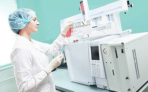 Female scientific researcher with lab coat, gloves and hair protection placing vial flask in gas chromatography instrument.