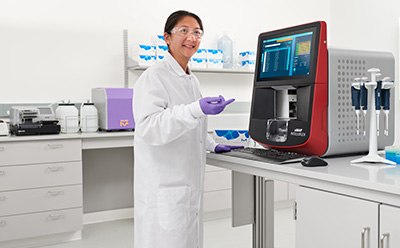 Female scientist in a lab with white coat, goggles, and gloves standing in front of a bench pointing to an xMAP® INTELLIFLEX® instrument that has pipettes and MIILIPLEX® multiplex immunoassay kits next to it.