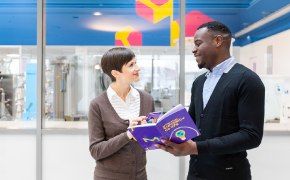 Employees discussing a book in a lab, with laboratory equipment visible in the background.