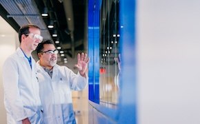 Two scientists in white lab coats are engaged in discussion while observing a digital display in a modern laboratory setting, with a focus on their gestures and the high-tech environment.