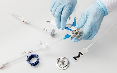 戴着蓝色手套的双手一丝不苟地组装或拆卸医疗或科学仪器。这些部件，包括管子、连接器以及塑料和金属制成的专用零件，散布在白色的表面上。其中一只手拿着一个有多个出口的部件，可能是用来连接管子或电线的。