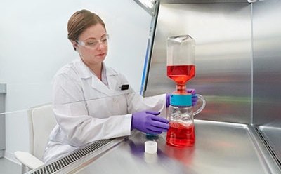 The image show a woman in a white lab coat, working with laboratory flask containing a red liquid, which is connected to another piece of equipment with blue liquid at the bottom.