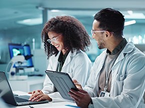 A professional setting featuring two individuals in lab coats engaged in a discussion while reviewing information on a tablet. The background is a soft green color, creating a clean and modern look. The image conveys a collaborative atmosphere in a scientific or healthcare environment.