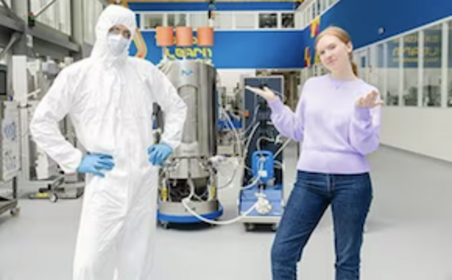 Two scientists, standing in a lab, a male in bunny suit, a female wearing casual clothes