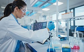 Dark-haired female lab worker wearing safety goggles, white lab coat and blue nitrile gloves prepares a solution of reagents. Glass vials, glass beakers and a computer screen are visible in the background.