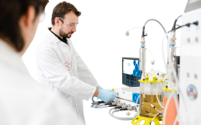 A scientist makes adjustments to a bioreactor while a second scientist watches from the foreground.