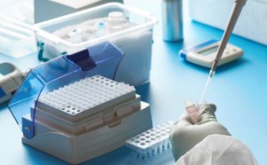 A laboratory scene showing a gloved hand using a pipette to transfer purified water into a sample tube, with various lab equipment in the background.