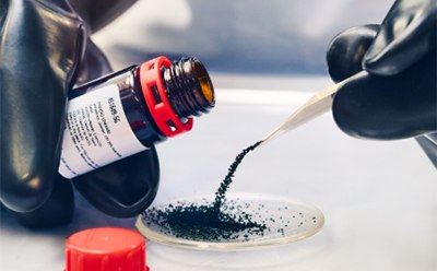 A person wearing black gloves carefully transfers a granular dark green or black powder from a labeled brown glass vial into a petri dish using metal tweezers in a laboratory setting. A red cap lies on the table nearby.