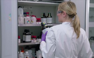 Shows the backside of a researcher wearing a white lab coat who is opening up a cabinet full of chemicals.